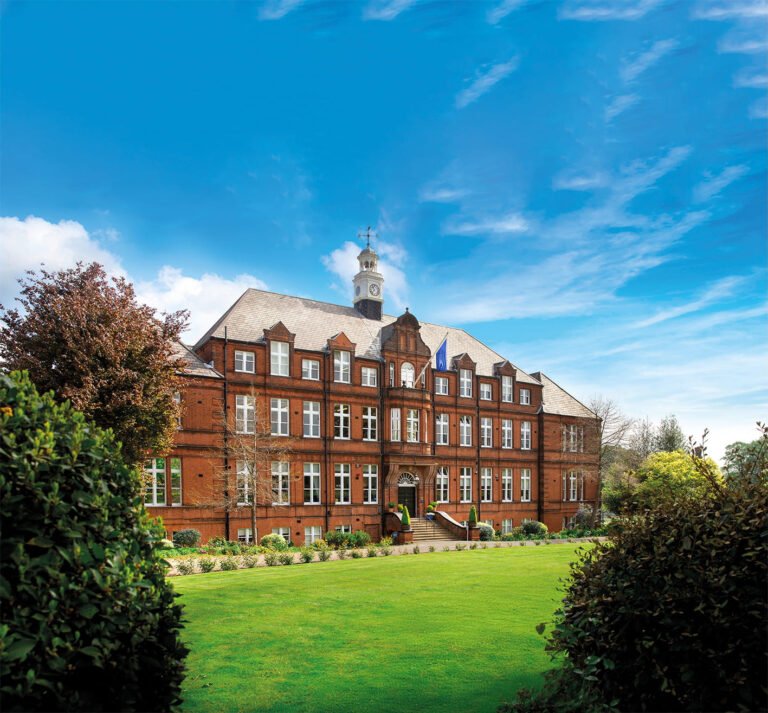 The grand red-brick exterior of Alleyn's School under a bright blue sky with a manicured green lawn in the foreground