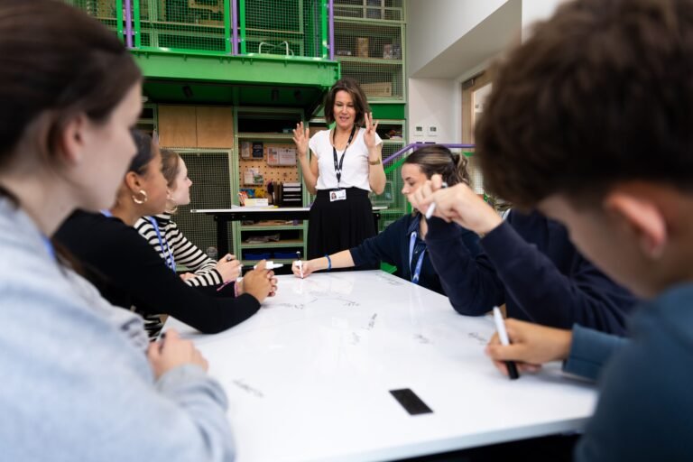 A teacher standing at the head of a large white table where several students are writing and brainstorming together in a modern classroom.