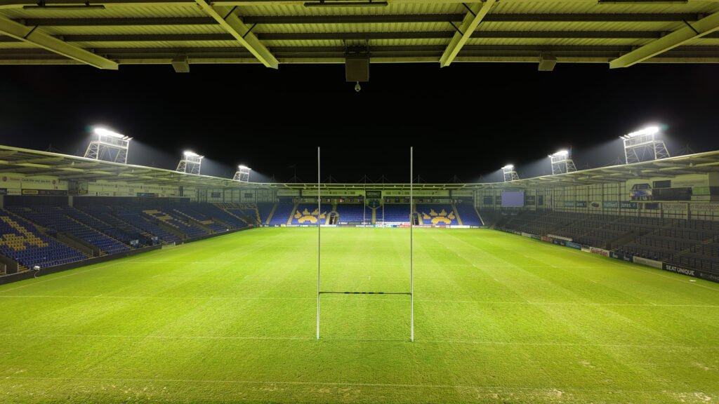 Wide-angle nighttime view behind the goalposts at the Warrington Wolves stadium, featuring the new LEDTURE LED floodlights and Protopixel dynamic control platform illuminating the pitch