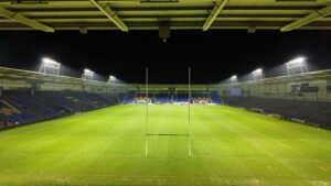 Wide-angle nighttime view behind the goalposts at the Warrington Wolves stadium, featuring the new LEDTURE LED floodlights and Protopixel dynamic control platform illuminating the pitch