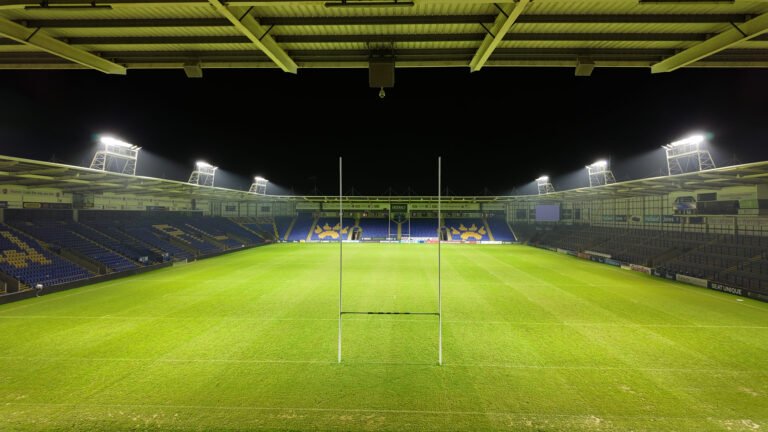 Wide-angle nighttime view behind the goalposts at the Warrington Wolves stadium, featuring the new LEDTURE LED floodlights and Protopixel dynamic control platform illuminating the pitch