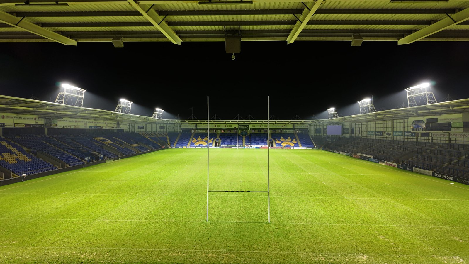 Wide-angle nighttime view behind the goalposts at the Warrington Wolves stadium, featuring the new LEDTURE LED floodlights and Protopixel dynamic control platform illuminating the pitch