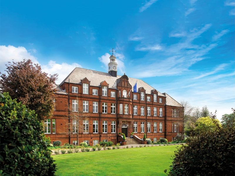 The grand red-brick exterior of Alleyn's School under a bright blue sky with a manicured green lawn in the foreground